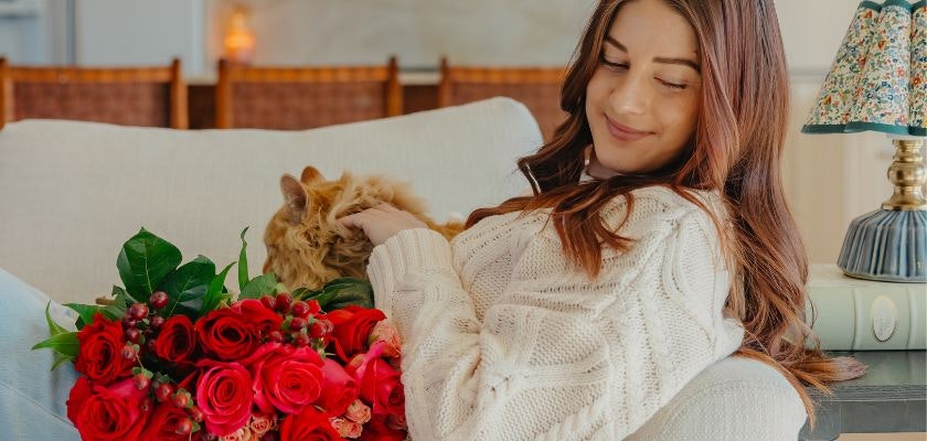 A woman in a cozy sweater enjoys a vibrant bouquet of red roses while sitting with her cat.