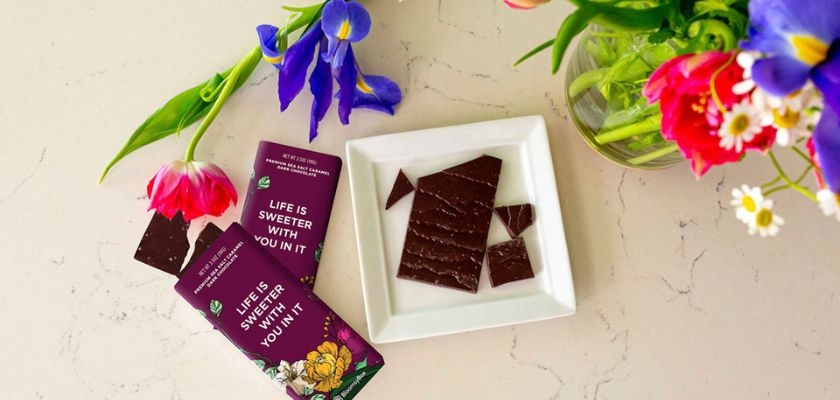 Delicious dark chocolate squares on a white plate next to vibrant flowers, showcasing a sweet treat.