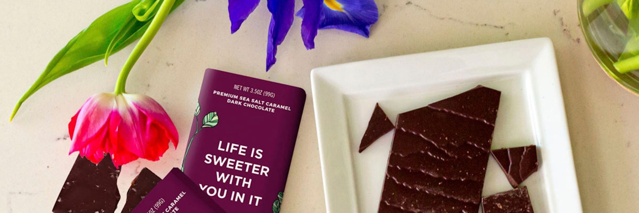 Vibrant tulip and iris flowers beside a bar of dark chocolate with a sweet slogan.