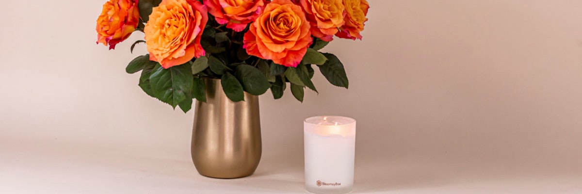 Vibrant orange roses in a sleek golden vase beside a glowing candle, creating a warm ambiance.