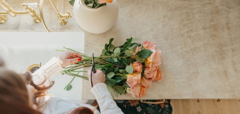 A person trimming peach roses with green leaves on a marble countertop for floral arrangements.
