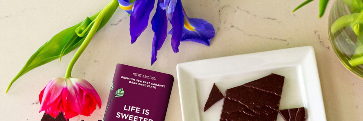A premium dark chocolate bar next to fresh colorful flowers on a marble surface.