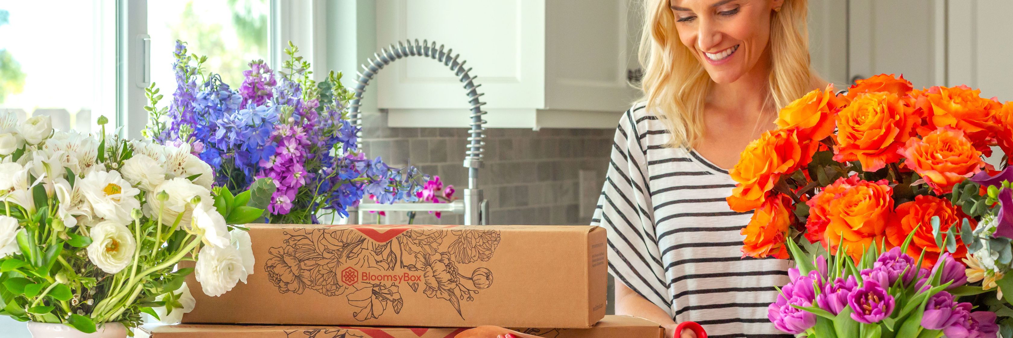 A cheerful woman unpacks colorful flower boxes in a bright kitchen, surrounded by vibrant blooms.