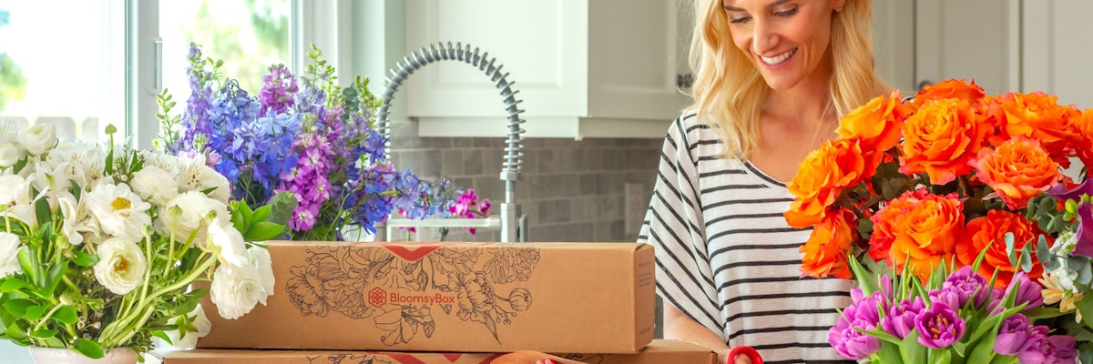 A cheerful woman unpacks colorful flower boxes in a bright kitchen, surrounded by vibrant blooms.