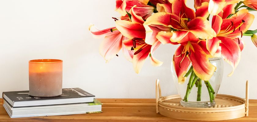 Vibrant orange and yellow lilies in a glass vase on a wooden table, beside a lit candle.