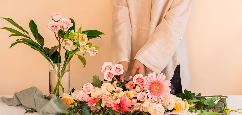 A person arranging a vibrant floral bouquet with pink roses and gerberas on a table.