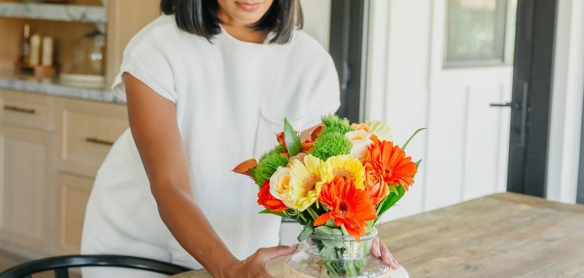 A woman arranging a vibrant floral bouquet with orange and yellow blooms on a rustic wooden table.
