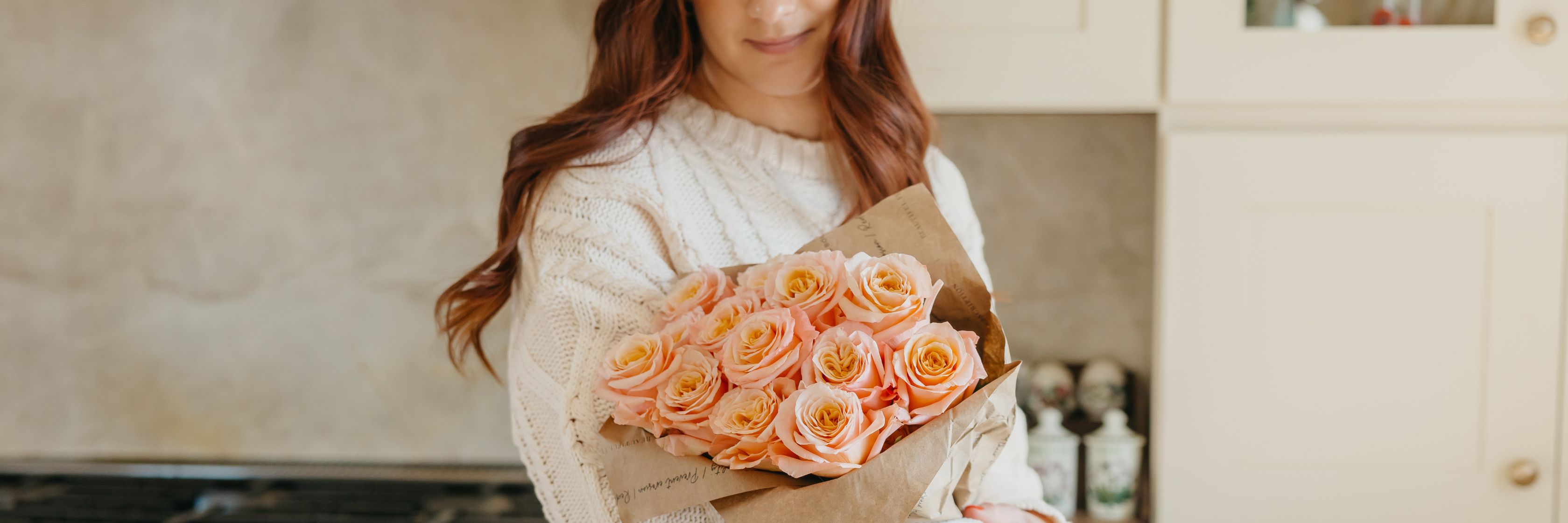 A woman wearing a cozy sweater holds a bouquet of soft pink roses wrapped in rustic paper.