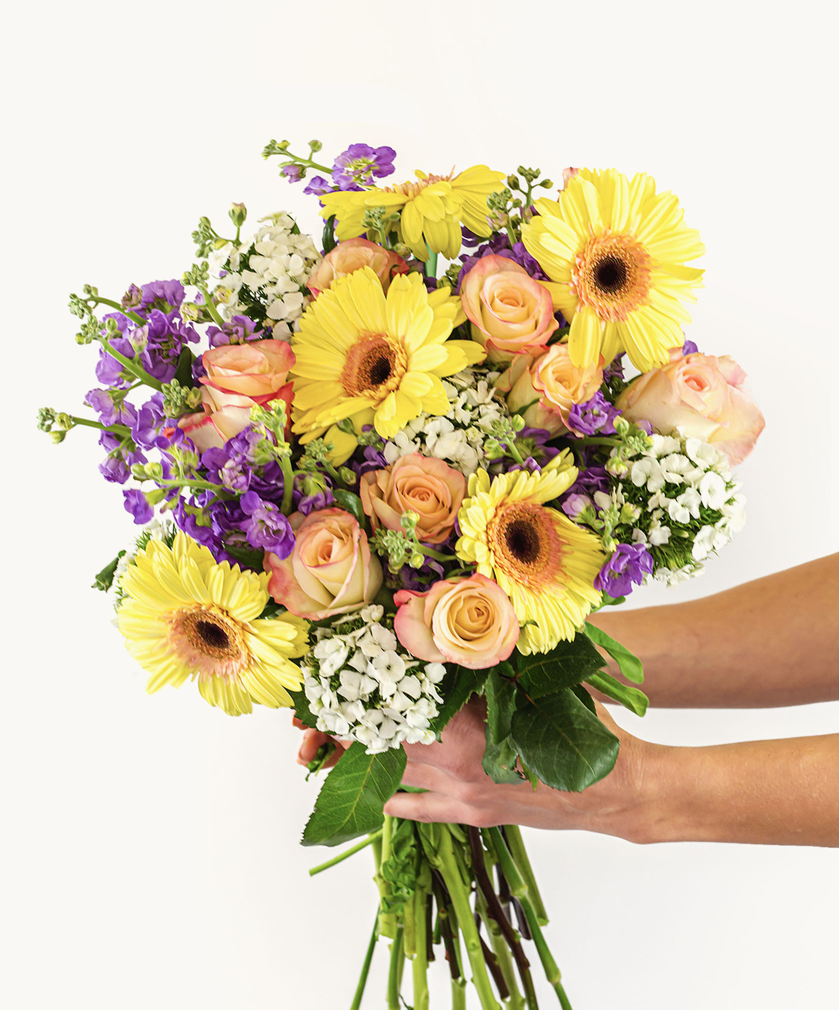 Vibrant bouquet featuring yellow gerbera daisies, pastel roses, and purple blooms, held gracefully.