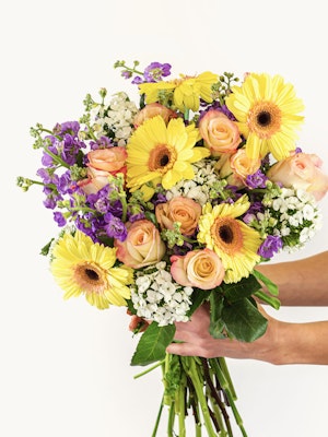 Vibrant bouquet featuring yellow gerbera daisies, pastel roses, and purple blooms, held gracefully.