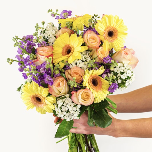 Vibrant bouquet featuring yellow gerbera daisies, pastel roses, and purple blooms, held gracefully.