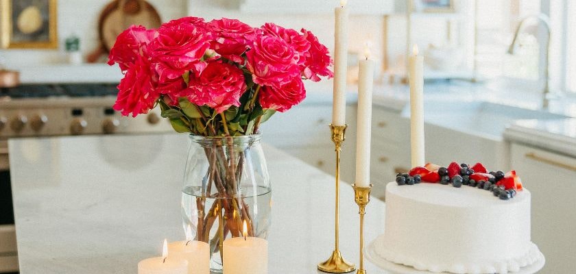 Vibrant pink roses in a glass vase, complemented by candles and a white cake on a marble table.