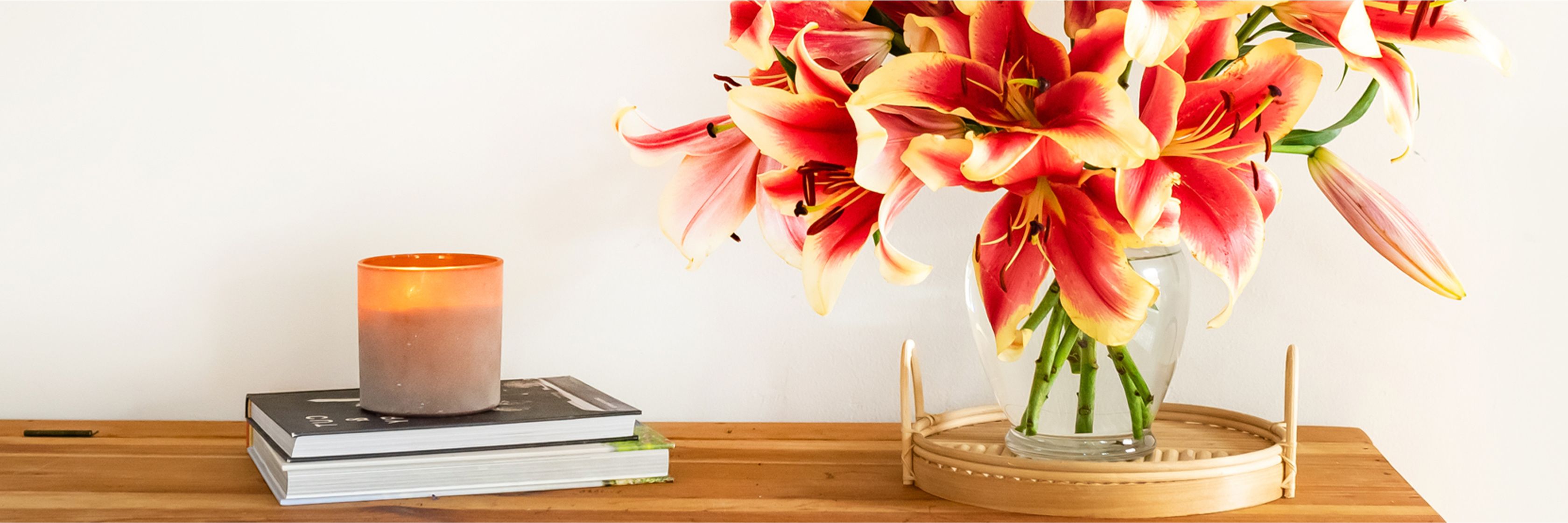 Vibrant lilies in a glass vase, paired with candles and stacked books on a wooden table.