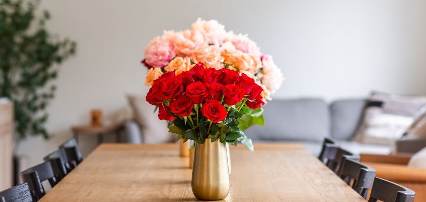Vibrant red roses and soft pink blooms arranged in a stylish gold vase on a wooden table.
