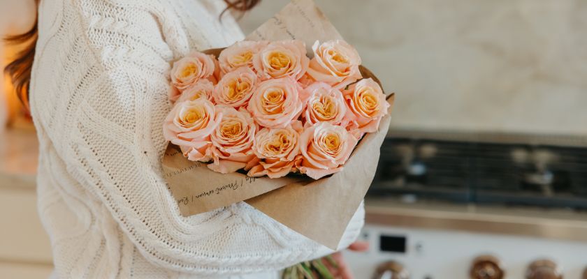 A lovely bouquet of peach roses wrapped in brown paper, held against a kitchen backdrop.