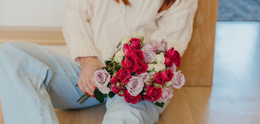 A woman in a cozy sweater holds a vibrant bouquet of pink and purple roses, sitting indoors.