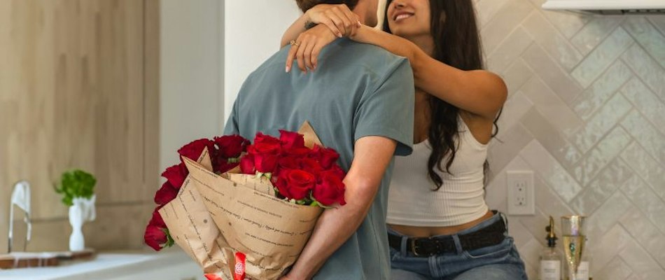 A couple sharing a romantic moment, man holding red rose bouquet behind his back.