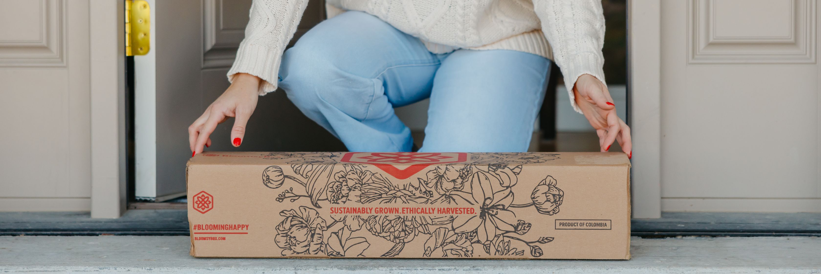 A woman in a cozy sweater receives a floral package at her doorstep, showcasing sustainable flowers.