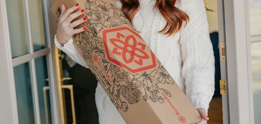 A woman in a cozy sweater is holding a floral delivery box from BloomsyBox.