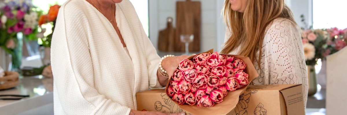 Two women exchanging a stunning bouquet of pink roses in a cozy, bright kitchen setting.