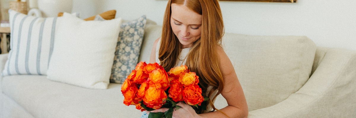 A woman with long red hair holds a vibrant bouquet of orange roses in her lap.
