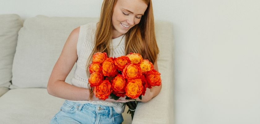 A young woman admires a vibrant bouquet of orange roses while sitting on a sofa.