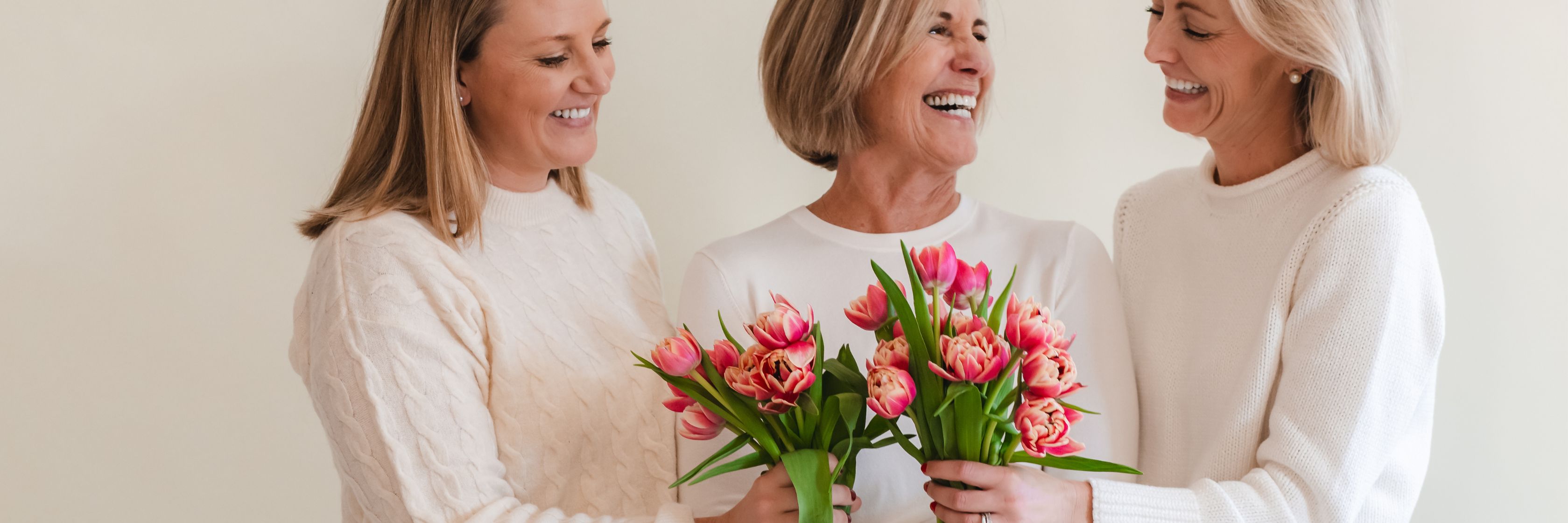 Three smiling women in cozy sweaters holding vibrant pink tulip bouquets, celebrating together.