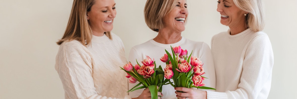 Three smiling women in cozy sweaters holding vibrant pink tulip bouquets, celebrating together.