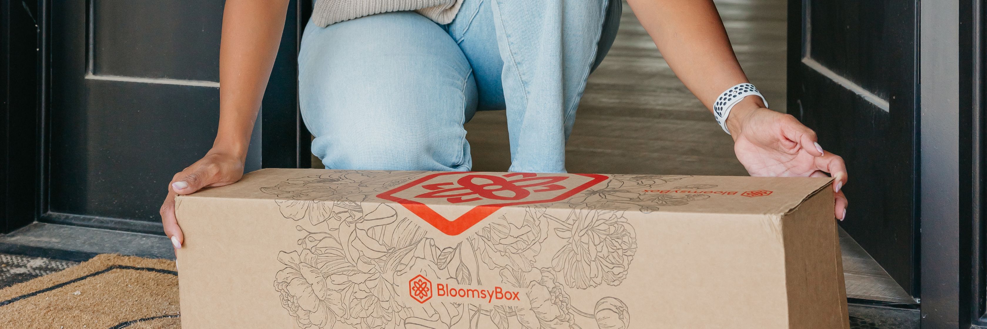 A person is delivering a BloomsyBox floral package at a home entrance.