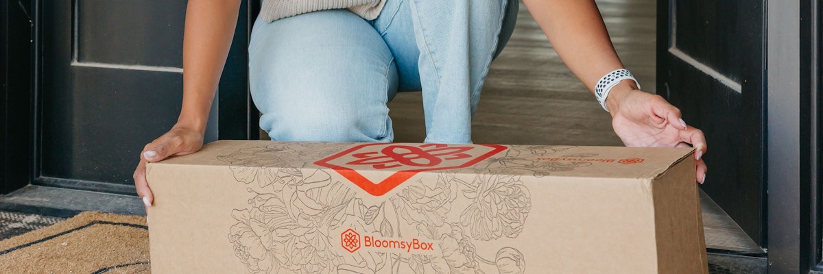 A person is delivering a BloomsyBox floral package at a home entrance.