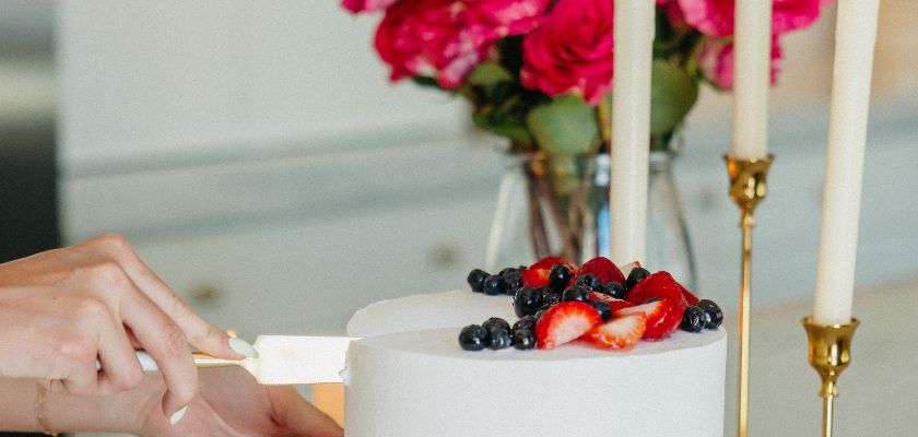 A delicious white cake topped with fresh berries, surrounded by a vibrant floral arrangement.