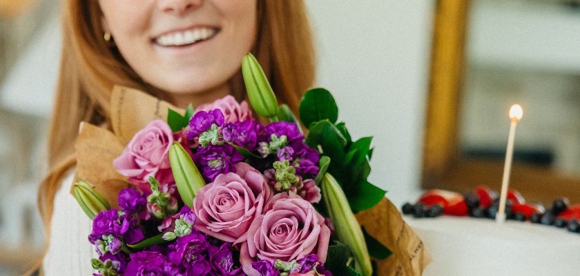 A joyful woman holding a vibrant bouquet of purple and pink flowers next to a cake.