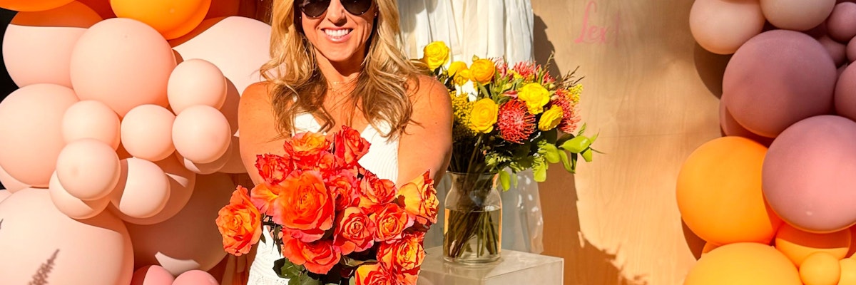 A cheerful woman in sunglasses holds a vibrant bouquet of orange roses amidst colorful balloons.