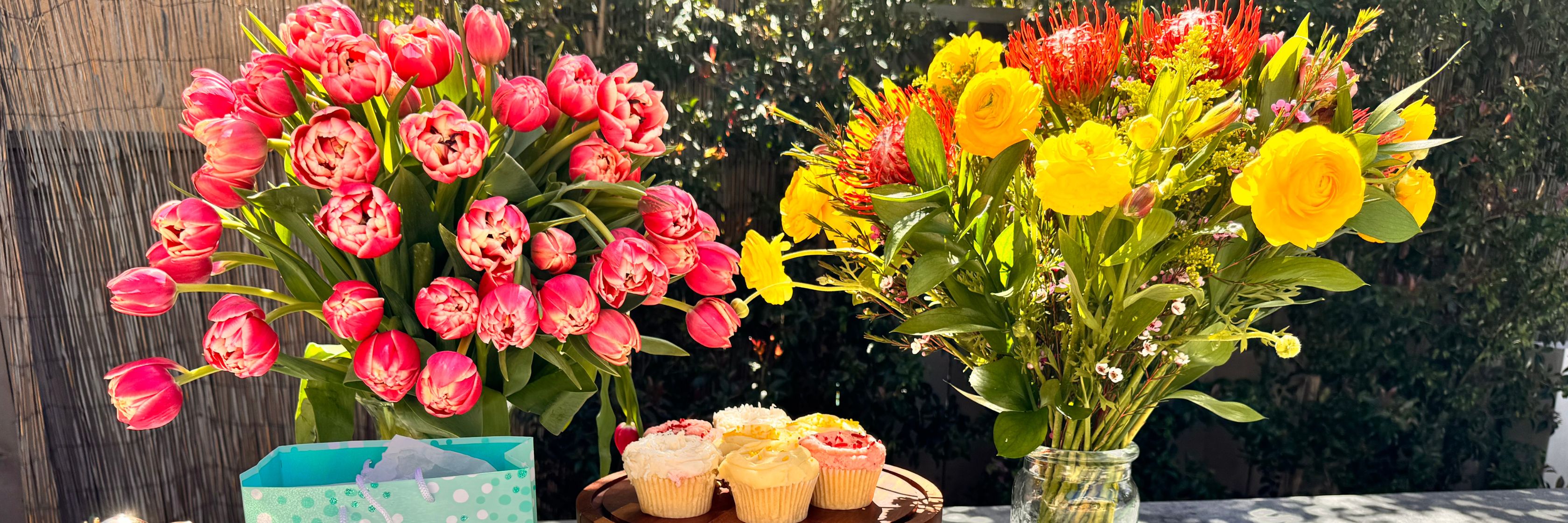 Brightly arranged bouquets of pink tulips and yellow roses beside colorful cupcakes.