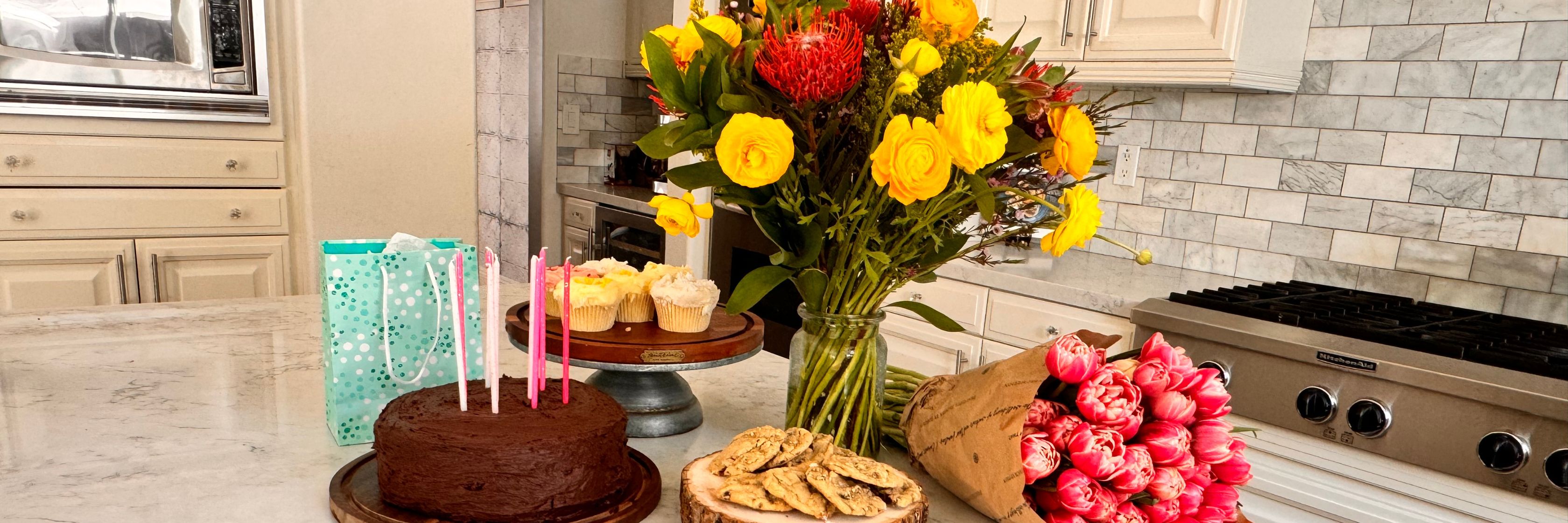 Colorful birthday celebration setting with a cake, flowers, and treats on a kitchen counter.