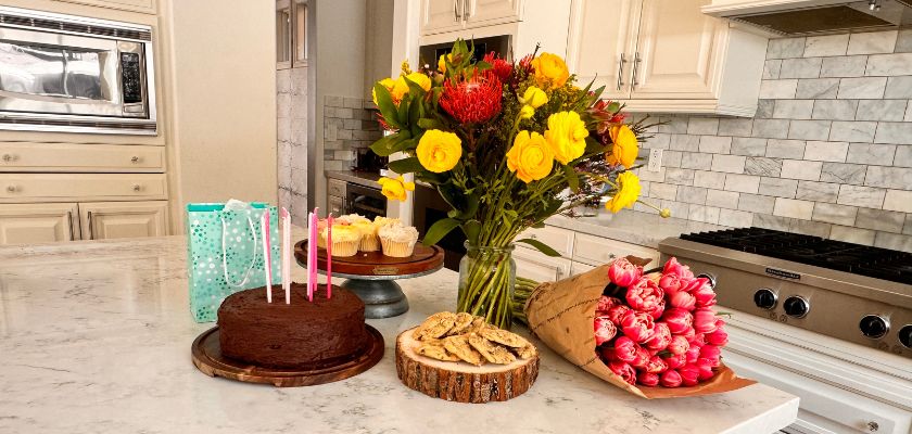 Colorful birthday celebration scene with a chocolate cake, flowers, and treats on a kitchen countertop.