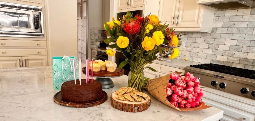 Colorful birthday celebration scene with a chocolate cake, flowers, and treats on a kitchen countertop.