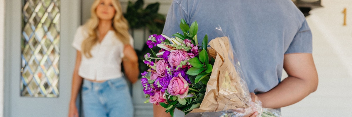 A person holding a colorful bouquet with purple and pink flowers, standing at a door.