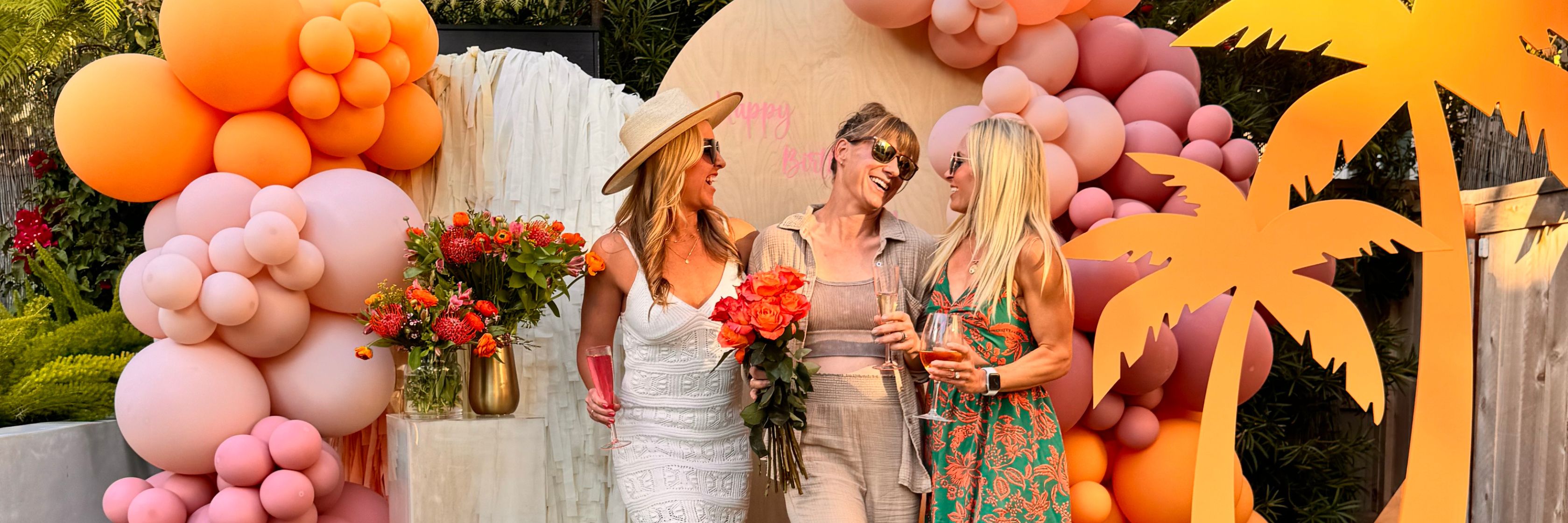 Three friends celebrating together in a festive setting with vibrant balloons and flowers.