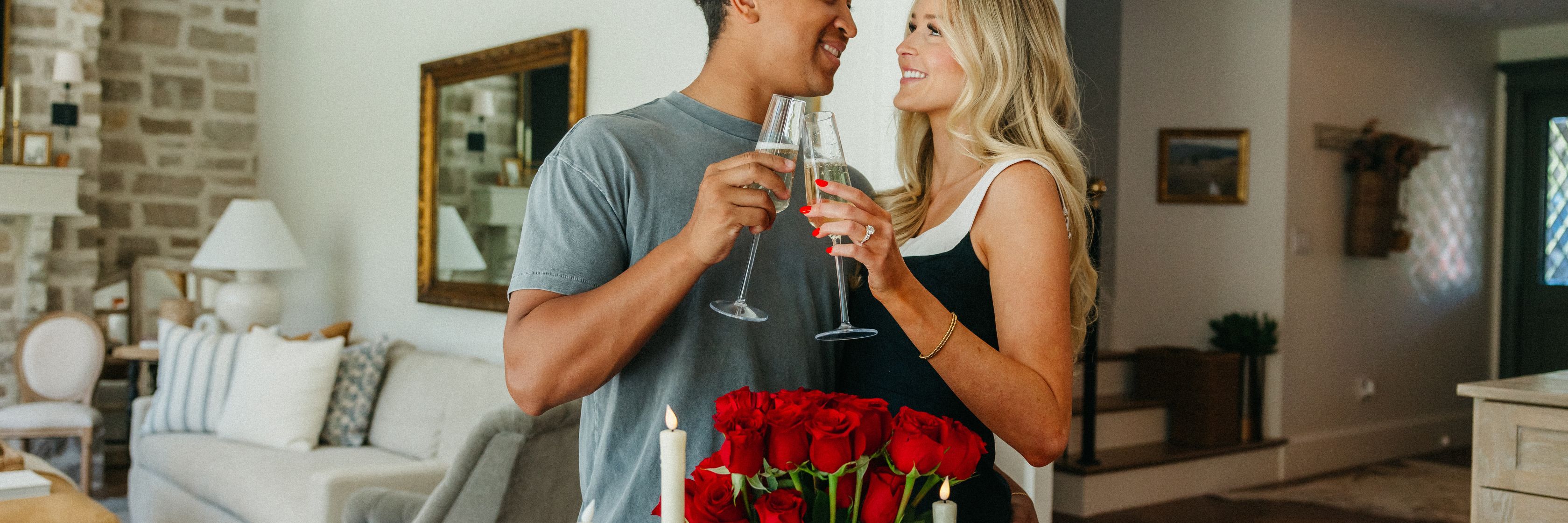 Couple celebrating with champagne and a bouquet of red roses in a cozy home setting.