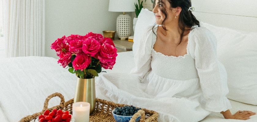 A stylish young woman sits on a bed with a vibrant rose bouquet and fresh berries.