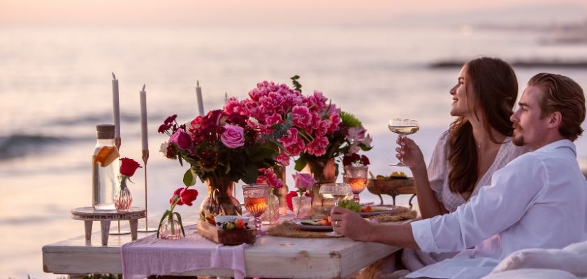 Couple enjoying a romantic beach dinner with pink roses and sunset views.