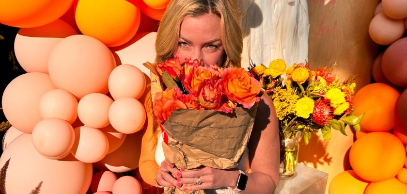 A woman joyfully holds a vibrant bouquet of orange flowers against a colorful balloon backdrop.