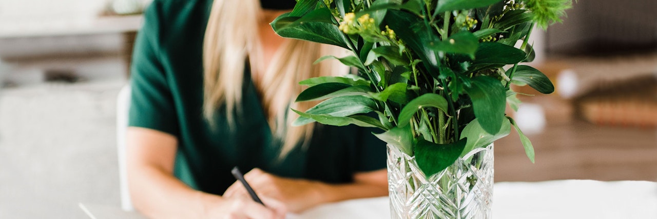 A woman in a green top writes notes beside a vibrant floral arrangement in a clear vase.