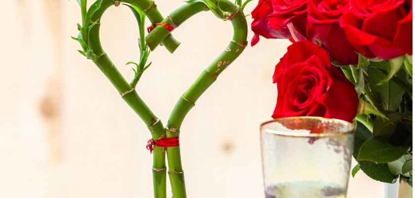 Heart-shaped bamboo arrangement beside vibrant red roses in a glass vase, symbolizing love.