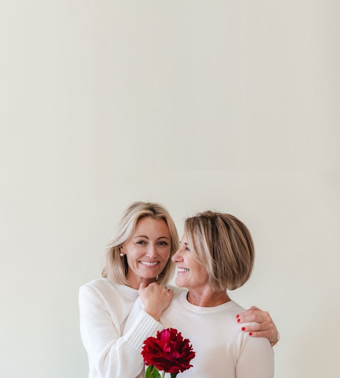 Two smiling women embrace, holding a vibrant red flower in a minimalist setting.