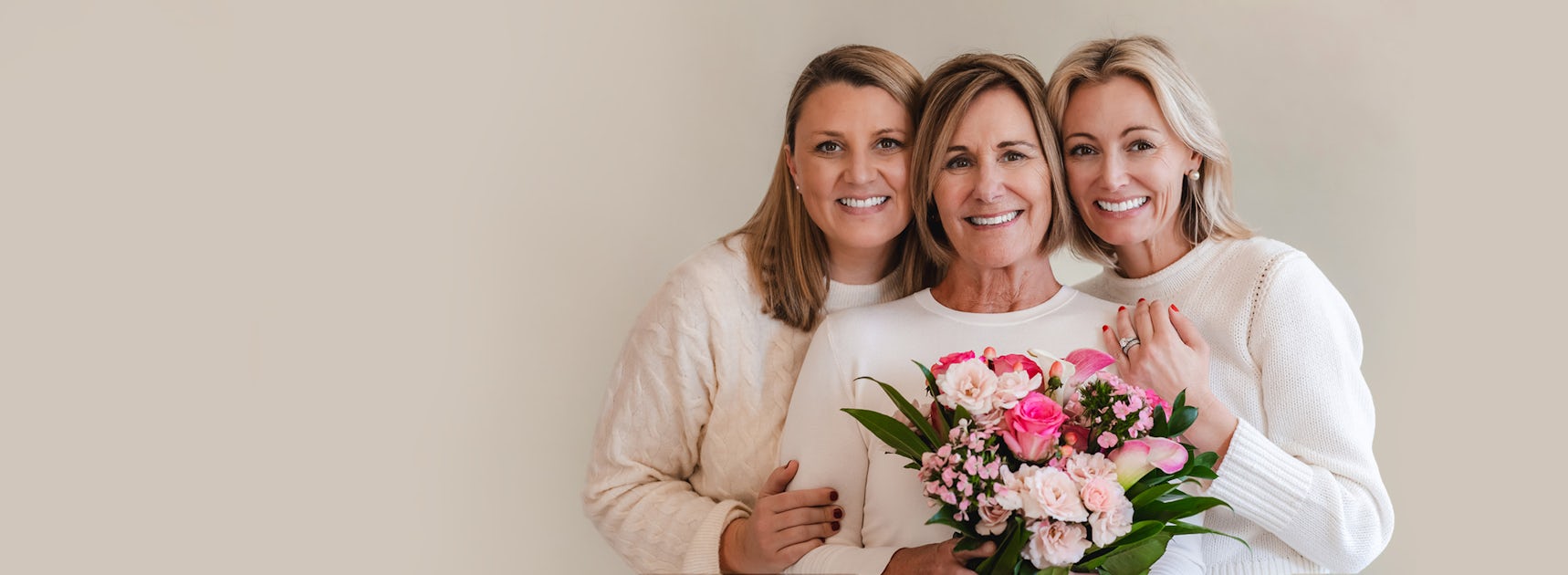 Three smiling women in cozy sweaters hold a vibrant floral bouquet together, celebrating friendship.