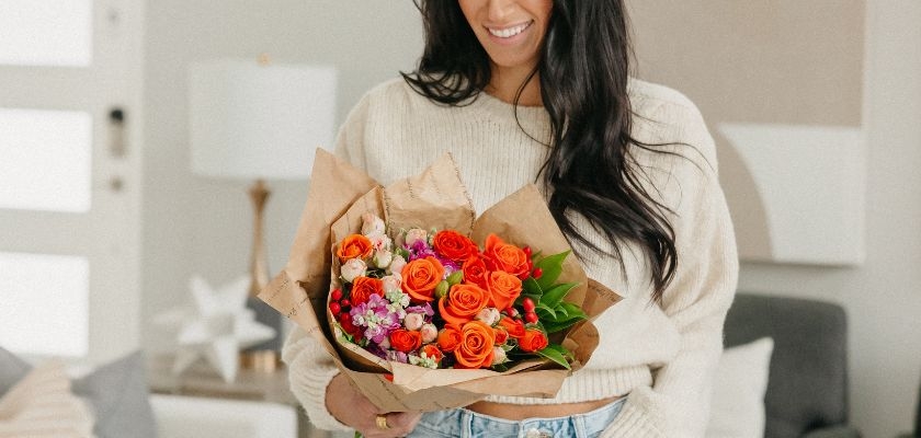 Cheerful woman holding a vibrant bouquet of orange roses and mixed blooms in kraft paper.