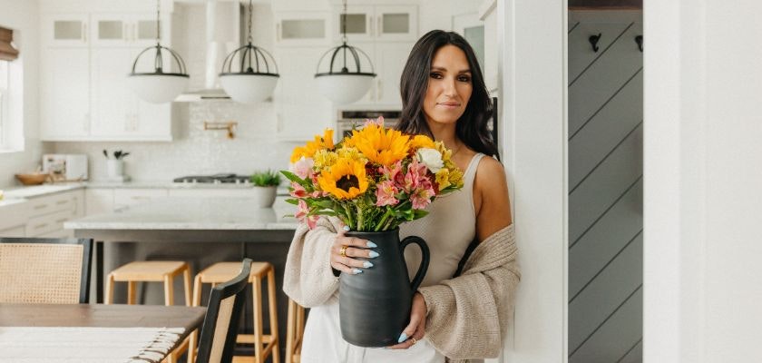 A woman holds a vibrant flower bouquet, featuring sunflowers and pink blooms, in her kitchen.