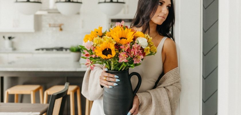A woman holds a vibrant bouquet of sunflowers and colorful blooms in a stylish pitcher.